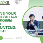A stressed business owner sitting at a cluttered desk overflowing with paper invoices, receipts, and spreadsheets, while a small, outdated calculator sits broken in the foreground. Behind them, a large wall chart shows a steep upward growth curve, signaling that the current basic bookkeeping system is failing. The image visually represents the 7 Signs Your Business has outgrown its current financial support structure.